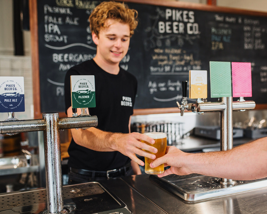 Attendant serving a beer at Pikes Beer Co. with a chalkboard menu in the background.