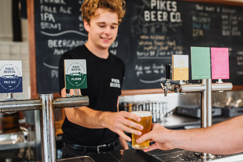 Attendant serving a beer at Pikes Beer Co. with a chalkboard menu in the background.