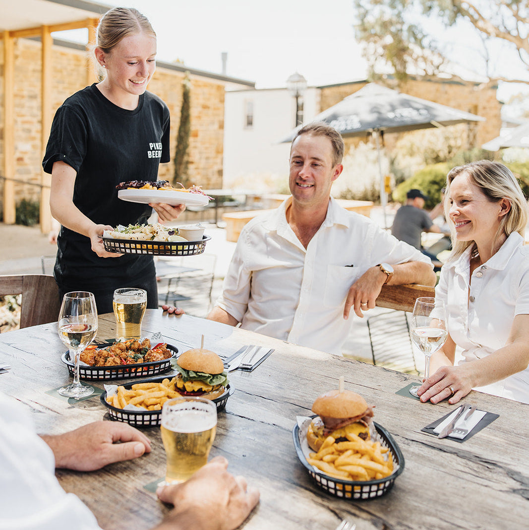 People enjoying a meal outdoors with a server, surrounded by trees and a building.