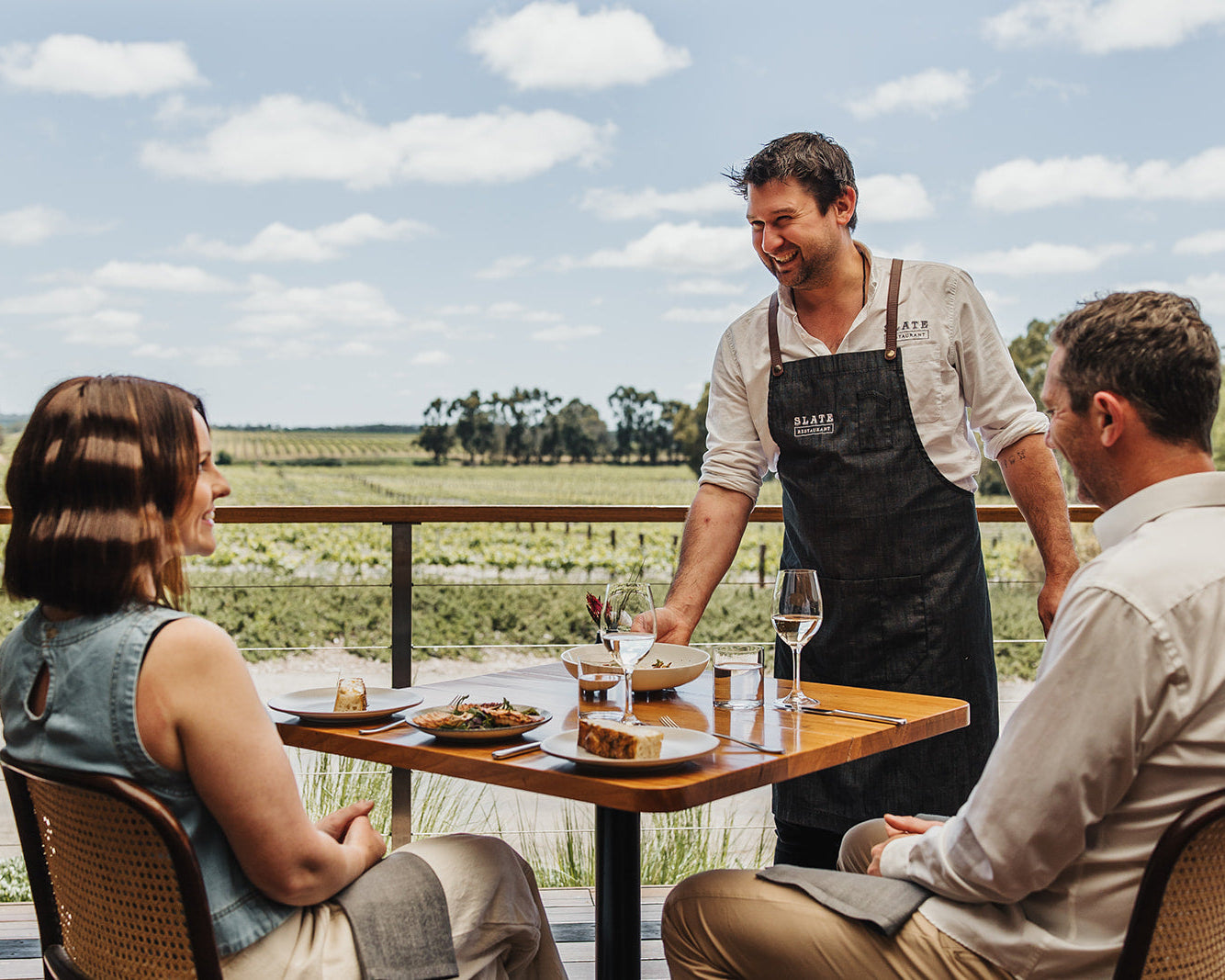 A smiling restaurant server wearing a black apron stands at a table with two seated customers outdoors on a patio overlooking a vineyard landscape under a blue sky.