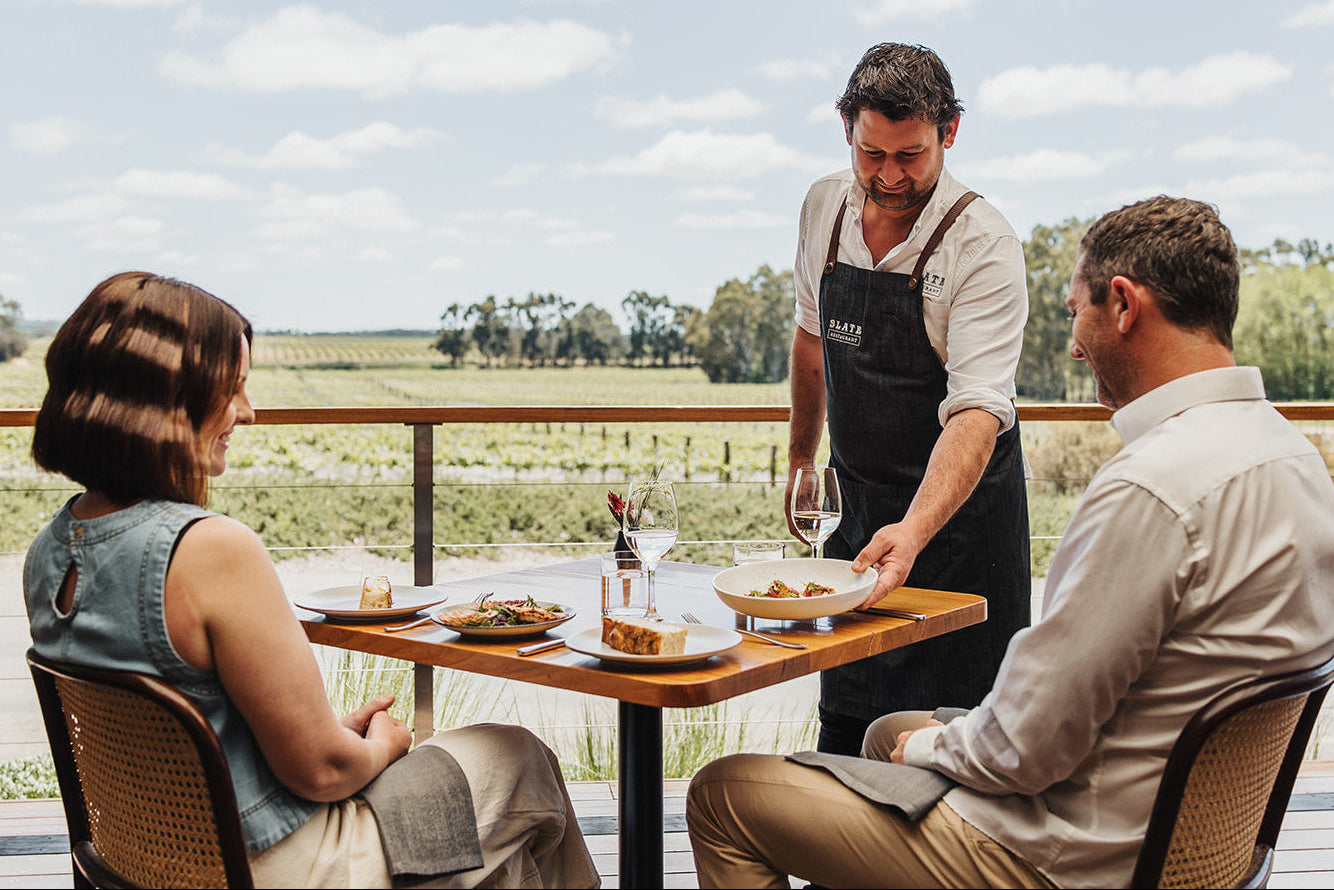 Waiter serving food to two guests on a balcony with a scenic view.