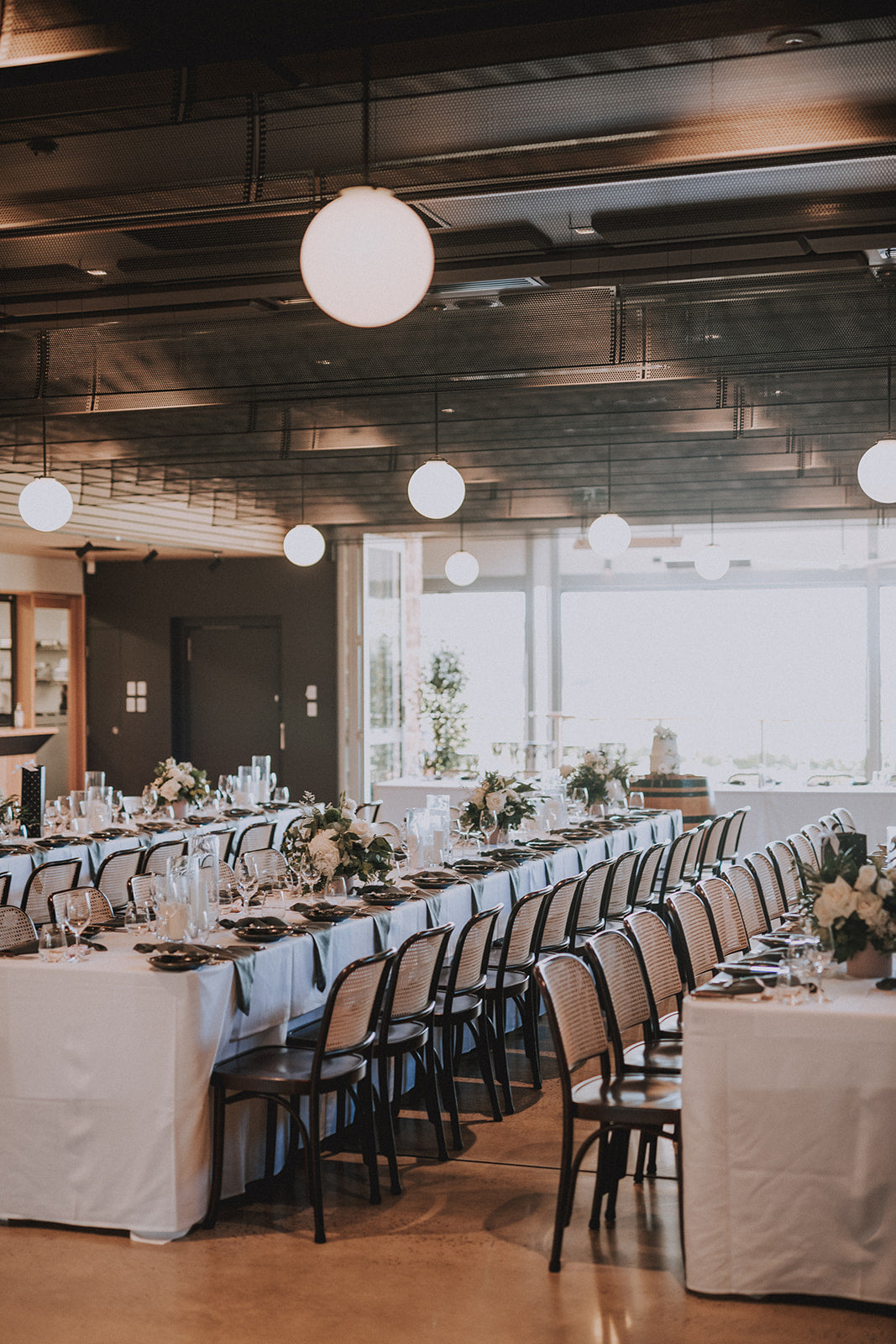 A long banquet table set in a modern event space with white tablecloths, black chairs, floral centerpieces, and hanging spherical pendant lights.