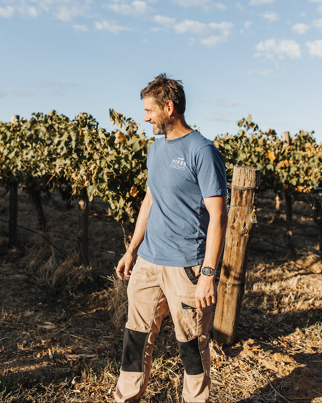 A man standing in a vineyard wearing a blue crewneck t-shirt with a white logo on the chest, khaki cargo pants, and knee pads, under a blue sky with light clouds.