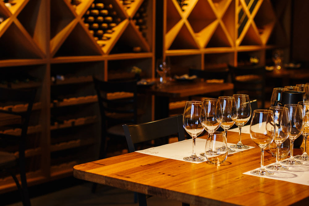 A warm, atmospheric restaurant or wine cellar interior with wooden tables, wine glasses, and geometric wood shelving filled with bottles in the background.