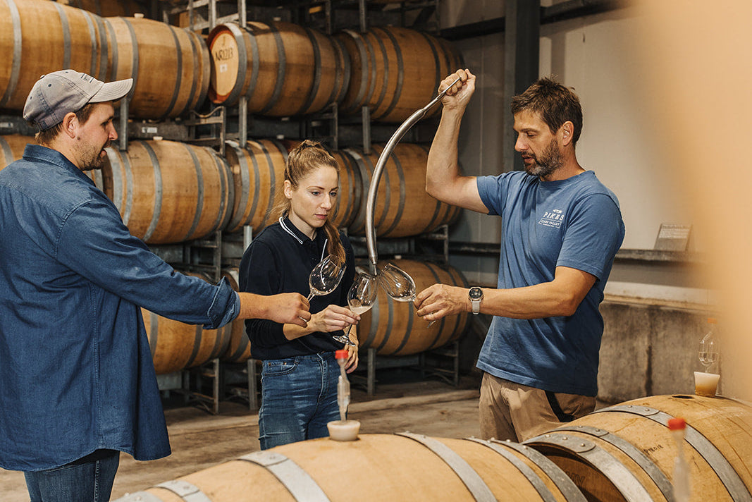 Three people in a wine cellar or distillery setting, with wooden barrels stacked in the background and one person pouring liquid from a large barrel into a small container held by the others.