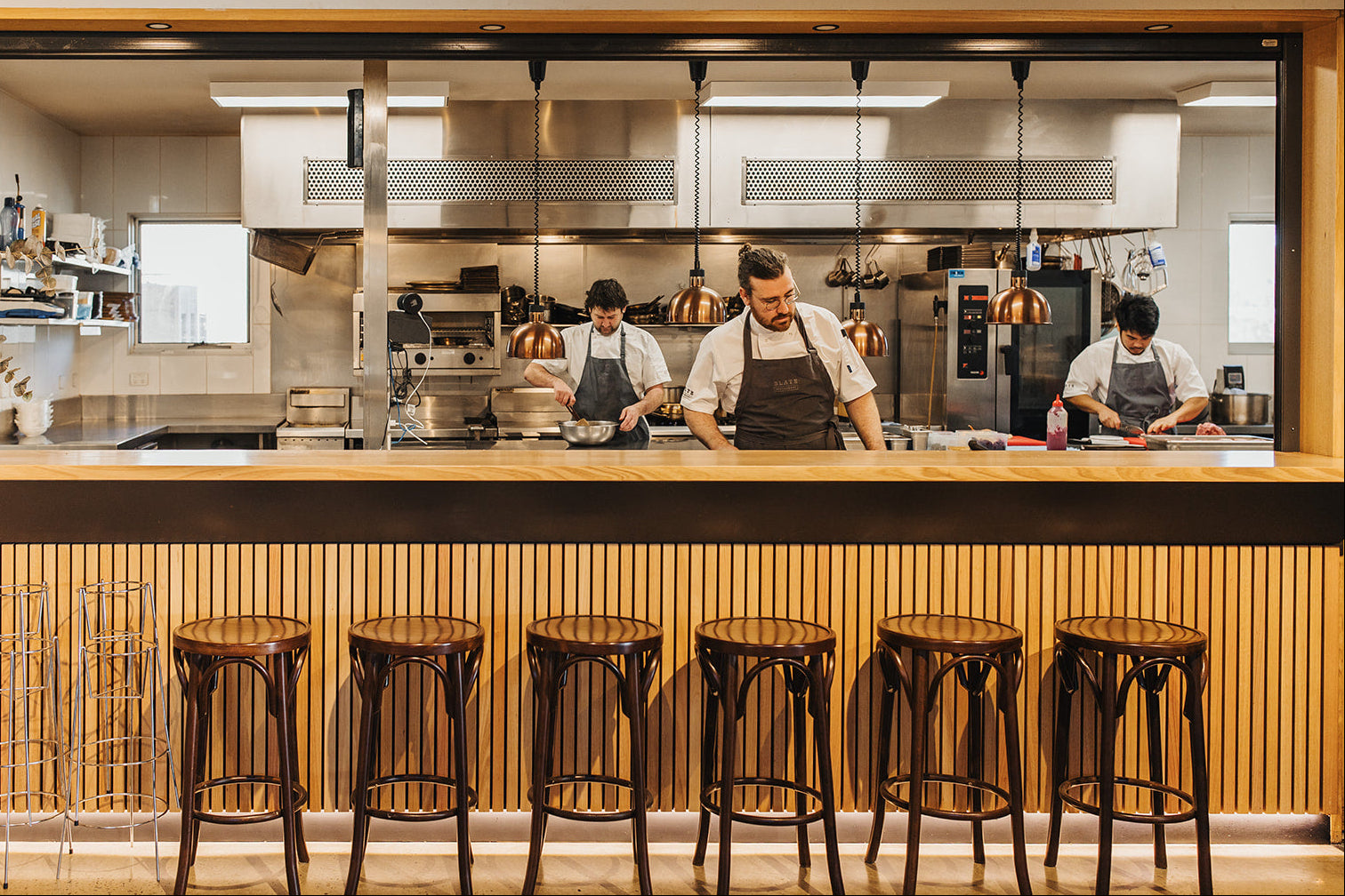 A restaurant interior with a wood counter and six dark metal bar stools in the foreground, overlooking a commercial kitchen where several chefs in aprons work behind a glass partition.